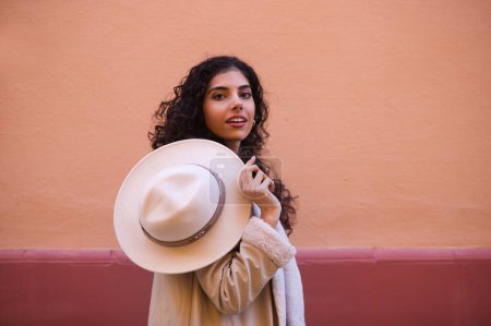 Téléchargez les photos : Jeune et belle femme brune hispanique aux cheveux bouclés joue avec un chapeau sur un fond rose. La femme joue à cache-cache derrière le chapeau en faisant différentes expressions drôles. - en image libre de droit