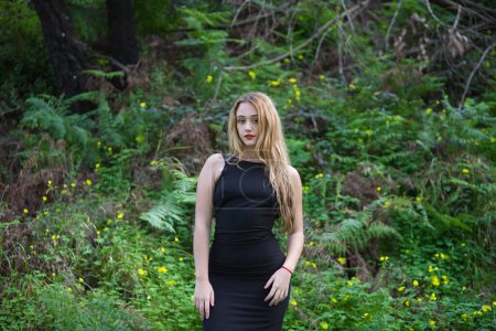 Foto de Joven, hermosa mujer rubia vestida de negro pasea por el bosque en diferentes posturas y expresiones. En el fondo helechos y flores amarillas. Expresiones conceptuales en la naturaleza. - Imagen libre de derechos