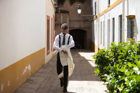 Photo for Handsome young man with a beard walking down the street takes off his white jacket. The man is on holiday in seville and walks through a typical neighbourhood of the city in spain. Holiday concept. - Royalty Free Image