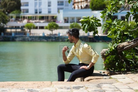 Foto de Joven guapo con barba y gafas de sol en la mano está sentado en las escaleras junto al río guadalquivir en Sevilla. En el fondo una higuera - Imagen libre de derechos