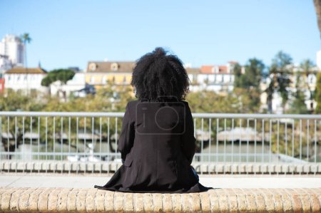 Foto de Joven, hermosa mujer negra con el pelo afro sentado en un banco mirando hacia el otro lado. Foto tomada por detrás. - Imagen libre de derechos