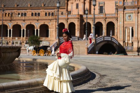 Photo for Young black woman dressed as a flamenco gypsy in a famous square in Seville, Spain. She is wearing a beige dress with ruffles and a red shawl and is standing in front of a fountain - Royalty Free Image