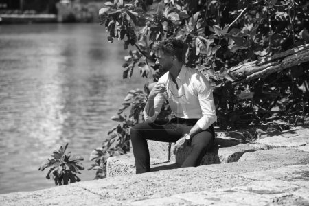 Foto de Joven guapo con barba y gafas de sol en la mano está sentado en las escaleras junto al río guadalquivir en Sevilla. Foto en blanco y negro. Monocromo - Imagen libre de derechos