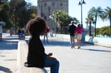 Foto de Joven, hermosa mujer negra con el pelo afro sentado en un banco frente al lado de la calle. Concepto de viaje y vacaciones. - Imagen libre de derechos
