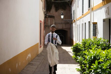 Photo for Handsome young man with a beard walking down the street takes off his white jacket. The man is on holiday in seville and walks through a typical neighbourhood of the city in spain. Holiday concept. - Royalty Free Image