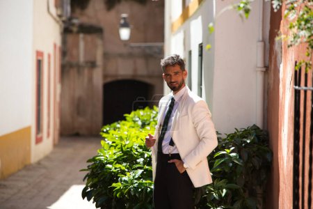 Hübscher junger Mann mit Bart, weißer Jacke und schwarzer Hose schlendert durch die Straßen der Innenstadt von Sevilla und posiert für Fotos wie ein Model. Urlaubs- und Geschäftsreisekonzept
