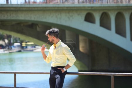 Foto de Un joven guapo con barba y gafas de sol en la mano se apoya en la barandilla del río guadalquivir en Sevilla. En la otra orilla está el famoso barrio de los artistas flamencos de Sevilla - Imagen libre de derechos