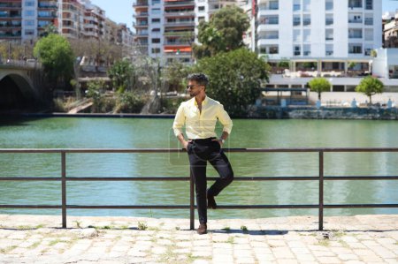 Foto de Joven guapo con barba y gafas de sol apoyado en la barandilla del río Guadalquivir en Sevilla. En la otra orilla está el famoso barrio de los artistas flamencos de Sevilla. - Imagen libre de derechos