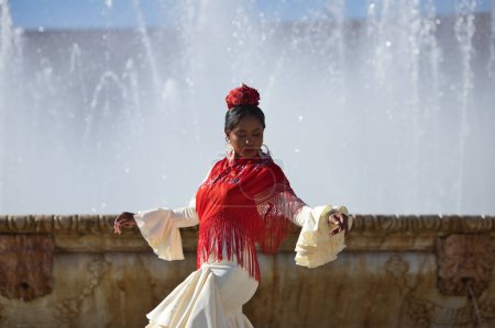 Téléchargez les photos : Jeune femme noire habillée en gitane flamenco sur une célèbre place de Séville, en Espagne. Elle porte une robe beige avec des volants et un châle rouge et se tient devant une fontaine - en image libre de droit