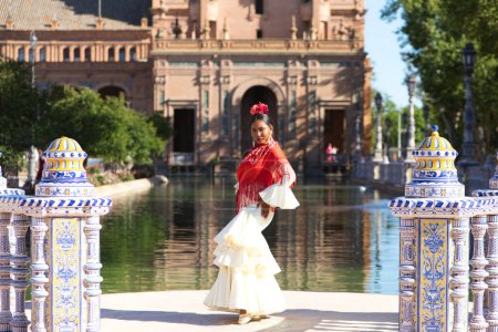 Téléchargez les photos : Jeune femme noire habillée en gitane flamenco sur une célèbre place de Séville, en Espagne. Elle porte une robe beige avec des volants et un châle rouge et est en face d'un canal sur la place - en image libre de droit