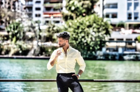 Foto de Un joven guapo con barba y gafas de sol en la mano se apoya en la barandilla del río guadalquivir en Sevilla. En la otra orilla está el famoso barrio de los artistas flamencos de Sevilla - Imagen libre de derechos