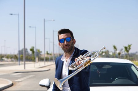 Photo for Handsome young man with beard, blue suit and sunglasses on his white car shows his trumpet in a happy attitude. The man is a musician. October 1st international day of music - Royalty Free Image