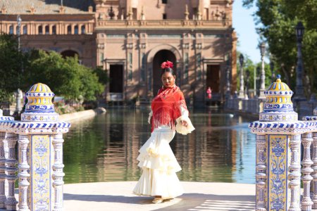Téléchargez les photos : Jeune femme noire habillée en gitane flamenco sur une célèbre place de Séville, en Espagne. Elle porte une robe beige avec des volants et un châle rouge et est en face d'un canal sur la place - en image libre de droit