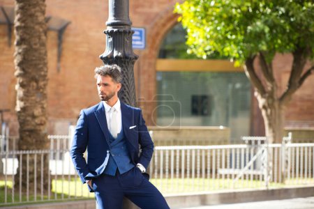 Téléchargez les photos : Beau jeune homme barbu et vêtu de costume bleu et cravate, appuyé contre un lampadaire, est en voyage d'affaires dans une ville européenne, Séville en Espagne - en image libre de droit
