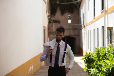 Photo for Handsome young man with a beard walking down the street and wearing a white jacket over his shoulder. The man is on holiday in seville and walks through a typical neighbourhood of the city in spain - Royalty Free Image