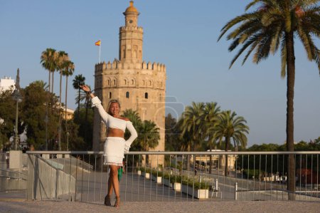 Photo for Beautiful young blonde woman from the United States is sightseeing in Seville, Spain. The girl is having fun doing different body positions and posing. In the background the monument the golden tower - Royalty Free Image