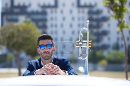 Photo for Handsome young man with beard, blue suit and sunglasses leaning on the car next to his trumpet. The man is a musician. October 1st international day of music. - Royalty Free Image