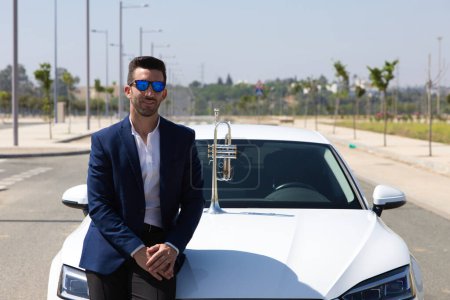 Photo for Handsome young man with beard, blue suit and sunglasses leaning on the car next to his trumpet. The man is a musician. October 1st international day of music. - Royalty Free Image