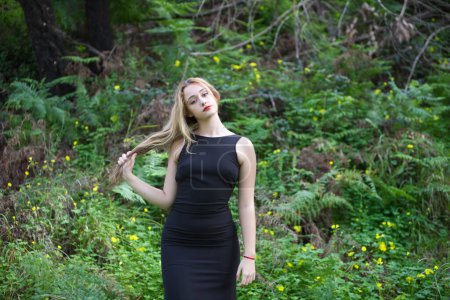 Junge, schöne blonde Frau in schwarz geht durch den Wald in verschiedenen Haltungen und Gesichtsausdrücken. Im Hintergrund Farne und gelbe Blumen. Konzeptausdrücke in der Natur.
