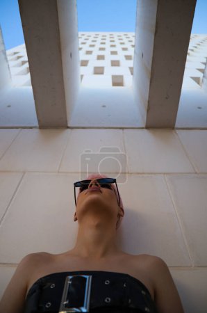 Photo for Young gay guy with pink hair and makeup and sunglasses is leaning on white wall making different expressions. From the top of the wall he gets daylight. Concept of equality and lgbtq rights - Royalty Free Image