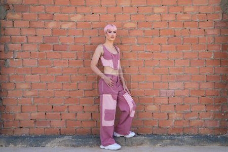 Young gay boy with pink hair and make-up leaning against a brick wall. The boy is dressed modernly in shades of pink. Concept of equality and LGBTQ rights