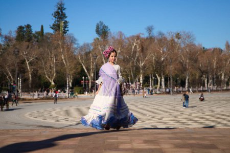 Foto de Una niña bailando flamenco vestida con un vestido beige con volantes y flecos morados en una famosa plaza de Sevilla, España. La chica tiene flores en la cabeza y el pelo en un moño. - Imagen libre de derechos