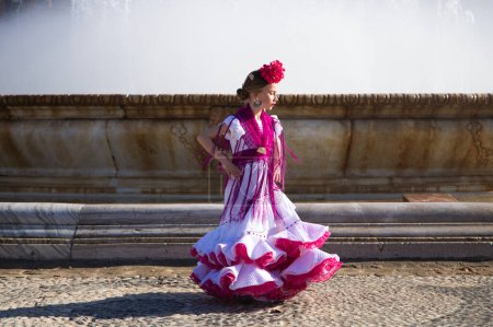 Foto de Una bonita niña bailando flamenco vestida con un vestido blanco con adornos rosas y flecos en una famosa plaza de Sevilla, España. La chica tiene una flor en el pelo. En el fondo una gran fuente - Imagen libre de derechos