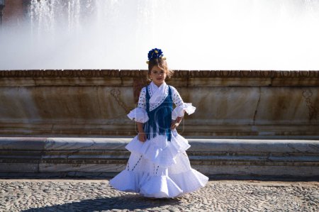 Foto de Una bonita niña bailando flamenco vestida con un vestido blanco con volantes y flecos azules en una famosa plaza de Sevilla, España. La chica tiene una flor en el pelo. En el fondo una gran fuente - Imagen libre de derechos
