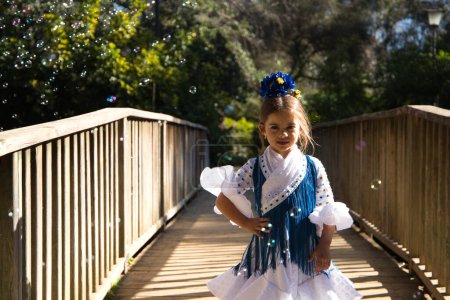 Foto de Una bonita niña bailando flamenco con un típico vestido gitano con volantes y flecos pasea sobre un puente de madera en un famoso parque de Sevilla, España. La chica posa entre las burbujas de jabón - Imagen libre de derechos