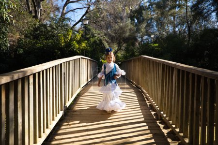 Foto de Una bonita niña bailando flamenco con un típico vestido gitano con volantes y flecos pasea sobre un puente de madera en un famoso parque de Sevilla, España. Su cabello está atado con una flor - Imagen libre de derechos