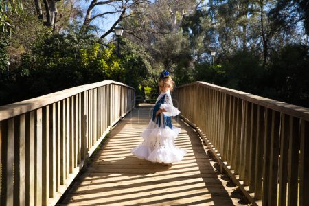 Foto de Una bonita niña bailando flamenco con un típico vestido gitano con volantes y flecos pasea sobre un puente de madera en un famoso parque de Sevilla, España. Su cabello está atado con una flor - Imagen libre de derechos