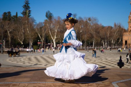 Foto de Una niña bailando flamenco vestida con un vestido blanco con volantes y flecos azules en una famosa plaza de Sevilla, España. La chica tiene flores en la cabeza. Flamenco, patrimonio cultural de la humanidad - Imagen libre de derechos