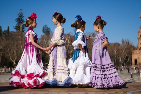 Foto de Cuatro niñas bailando flamenco vestidas con el típico traje flamenco se hablan en una famosa plaza de sevilla, España. Al fondo una antigua torre cuadrada. - Imagen libre de derechos