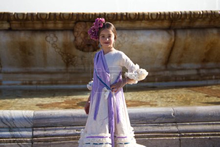 Foto de Una bonita niña bailando flamenco vestida con un vestido beige con volantes y flecos morados en una famosa plaza de Sevilla, España. La chica tiene una flor en el pelo. En el fondo una gran fuente - Imagen libre de derechos