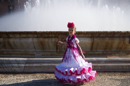Foto de Una bonita niña bailando flamenco vestida con un vestido blanco con adornos rosas y flecos en una famosa plaza de Sevilla, España. La chica tiene una flor en el pelo. En el fondo una gran fuente - Imagen libre de derechos