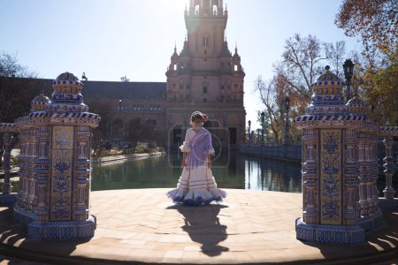 Una chica guapa bailando flamenco con un vestido de volantes y flecos en una famosa plaza de Sevilla, España. La chica lleva una flor en el pelo y le da la espalda. Al fondo una torre