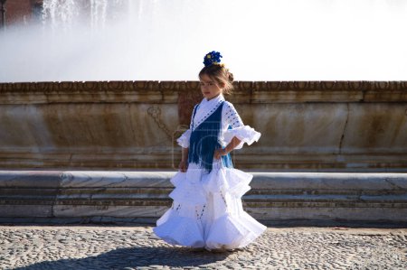 Foto de Una bonita niña bailando flamenco vestida con un vestido blanco con volantes y flecos azules en una famosa plaza de Sevilla, España. La chica tiene una flor en el pelo. En el fondo una gran fuente - Imagen libre de derechos