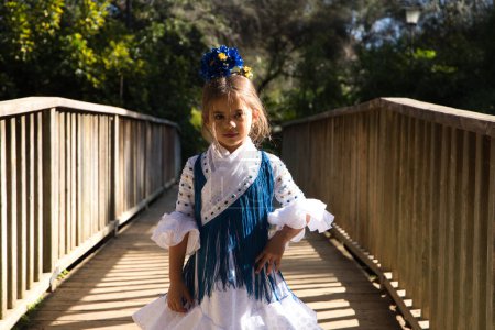 Foto de Retrato de una bonita niña bailando flamenco en un vestido con volantes y flecos típicos de los gitanos, caminando sobre un puente de madera en un famoso parque de Sevilla, España. Su cabello está atado con una flor - Imagen libre de derechos