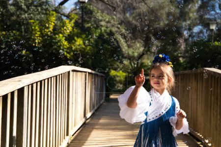 Foto de Una bonita niña bailando flamenco con un típico vestido gitano con volantes y flecos pasea sobre un puente de madera en un famoso parque de Sevilla, España. La chica juega con burbujas de jabón. - Imagen libre de derechos