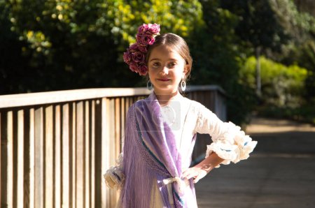 Foto de Retrato de una bonita niña bailando flamenco en un vestido con volantes y flecos típicos de los gitanos, caminando sobre un puente de madera en un famoso parque de Sevilla, España. Su cabello está atado con una flor - Imagen libre de derechos