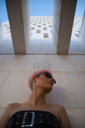 Photo for Young gay guy with pink hair and makeup and sunglasses is leaning on white wall making different expressions. From the top of the wall he gets daylight. Concept of equality and lgbtq rights - Royalty Free Image