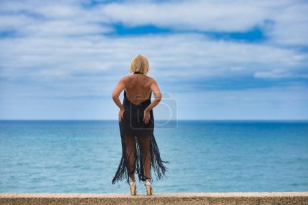 Mujer madura, rubia, hermosa, vestida con un elegante vestido negro mira con anhelo el horizonte sobre el océano atlántico en Cádiz, España. La mujer está triste y nostálgica. Foto tomada por detrás