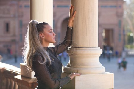 Hübsche junge blonde Frau mit Pferdeschwanz im Haar lehnt an einer Marmorsäule auf der Plaza de espana in Sevilla. Das Mädchen macht unterschiedliche Mienen. Tourismus- und Urlaubskonzept