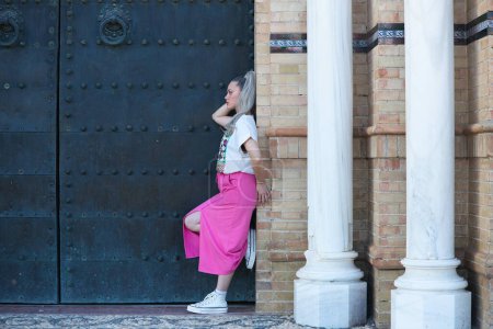 Beautiful young blonde woman leaning on the doorjamb of a large door of a monumental building in the city of Seville. The girl leans on one leg and touches her head. The girl is dressed casually
