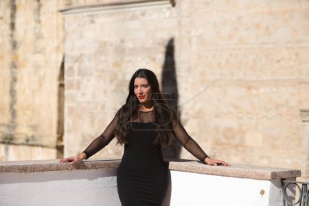 Téléchargez les photos : Belle jeune femme brune espagnole avec maquillage et vêtue d'une élégante robe noire. La femme s'appuie sur la balustrade. Concept beauté et mode - en image libre de droit