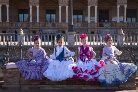 Foto de Cuatro niñas bailando flamenco vestidas con el típico traje flamenco sentadas en un banco en una famosa plaza de Sevilla, España. En el fondo los arcos y columnas de la famosa plaza - Imagen libre de derechos