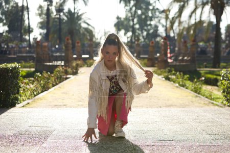 Young beautiful blonde woman with a ponytail in her hair wearing a pink skirt and fringed jacket rests one knee, one hand on the ground and with a defiant face looks at the camera. The girl is poised