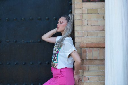 Beautiful young blonde woman leaning on the doorjamb of a large door of a monumental building in the city of Seville. The girl leans on one leg and touches her head. The girl is dressed casually