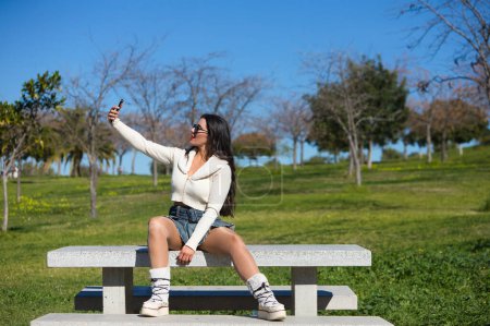 Young and beautiful Spanish brunette woman with sunglasses sitting on a park bench. The girl is casually dressed and takes a selfie with her mobile phone while enjoying the sunny spring day