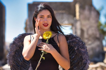 A young, beautiful, dark-haired latin woman with black angel wings in a ruined city clutches a sunflower in her hands. The woman is alone in the rubble and makes different sad and happy expressions
